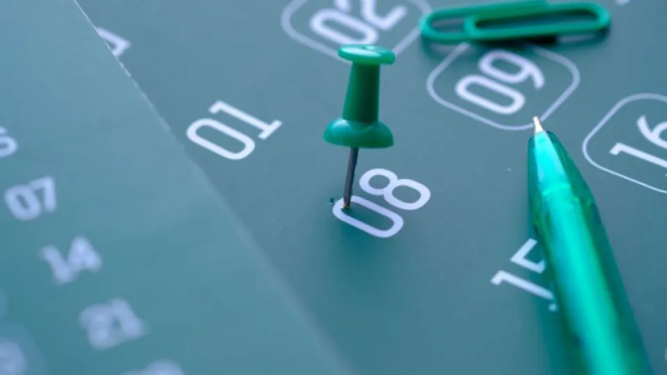 Close-up de um calendário com um alfinete verde marcado no dia 8, acompanhado de uma caneta e um clip também verdes.