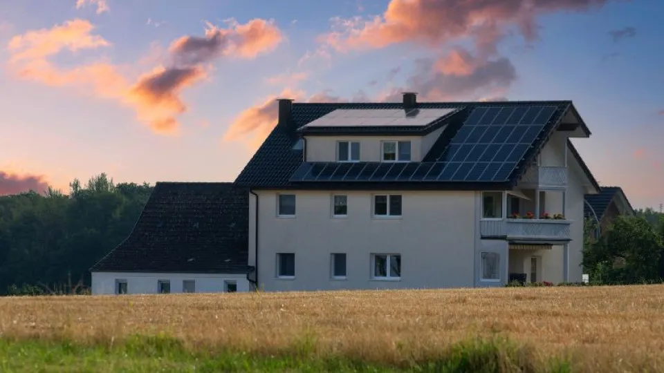 Casa moderna com painéis solares no telhado, situada numa área rural com campo de trigo dourado em primeiro plano e floresta ao fundo, ao entardecer com céu colorido em tons de rosa e azul.